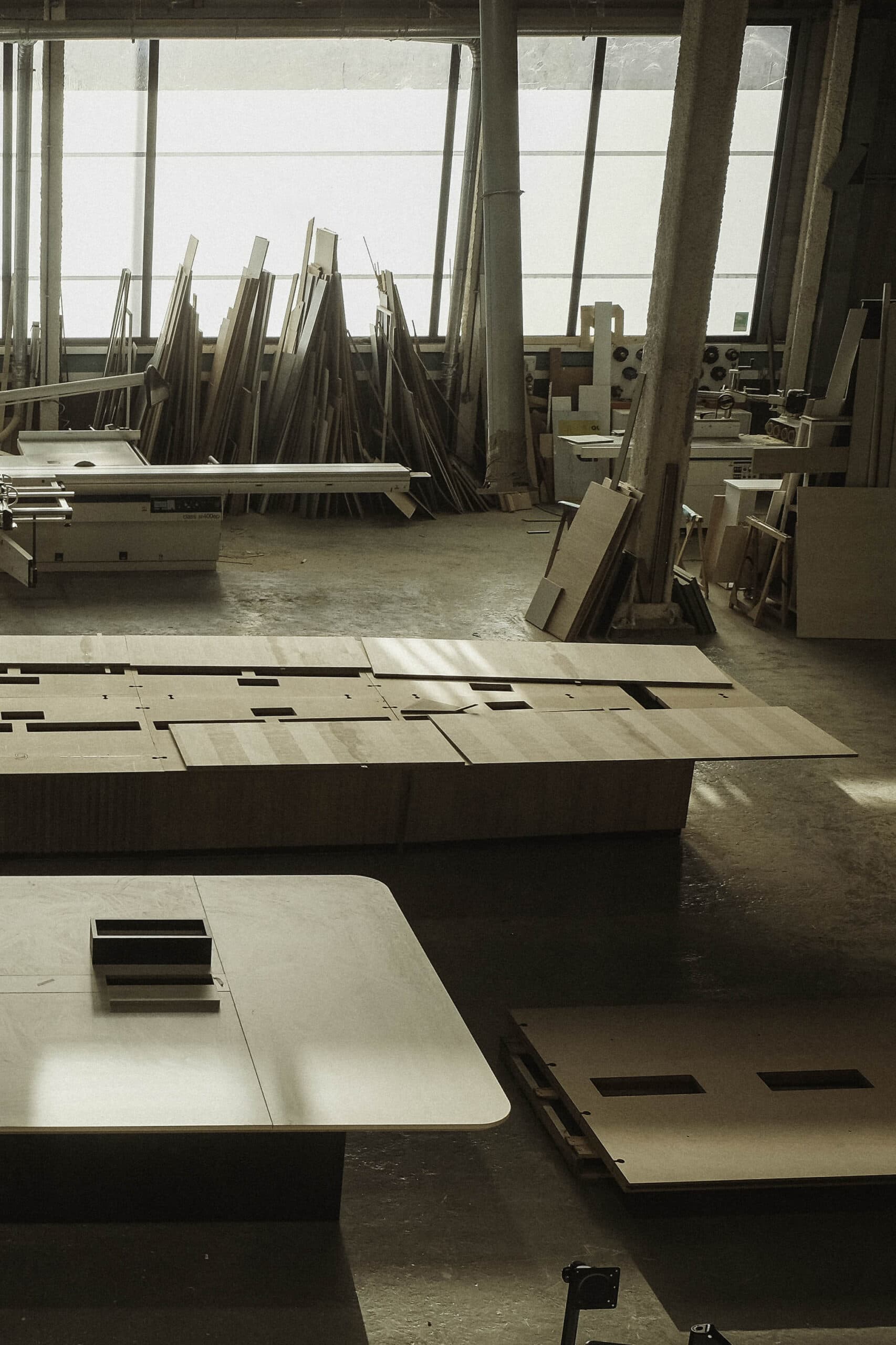 Dimly lit furniture workshop with large wooden panels, machinery, stacked timber, and unfinished pieces arranged across the floor.
