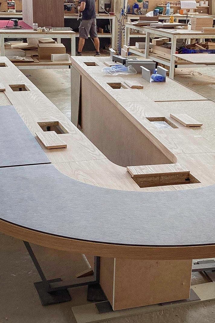 Curved custom furniture structure under construction in a busy workshop, with cut-outs, wood panels, tools, and materials visible.