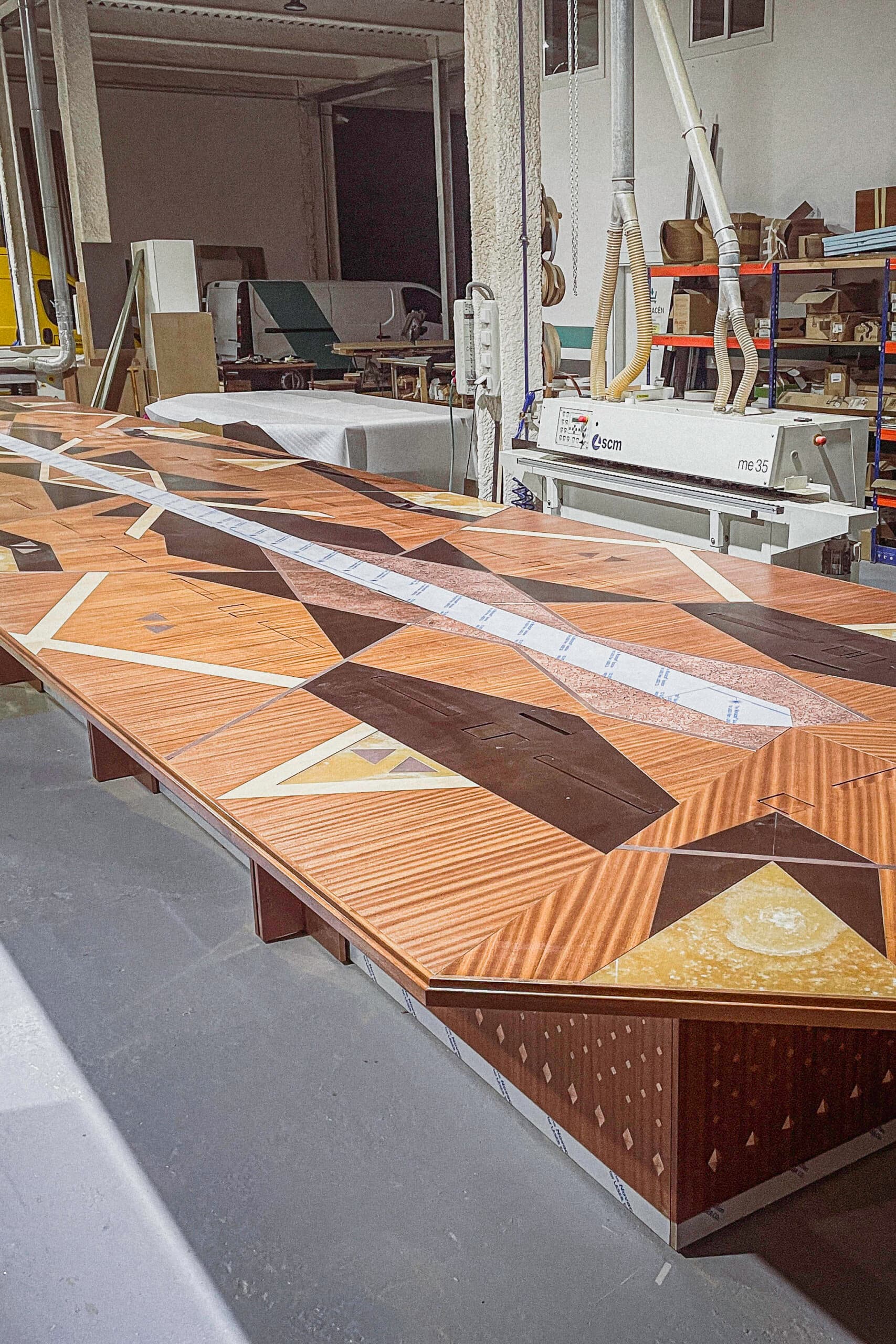 Large decorative wood-veneer surface being assembled in a workshop, featuring geometric inlays in varied tones and patterns.