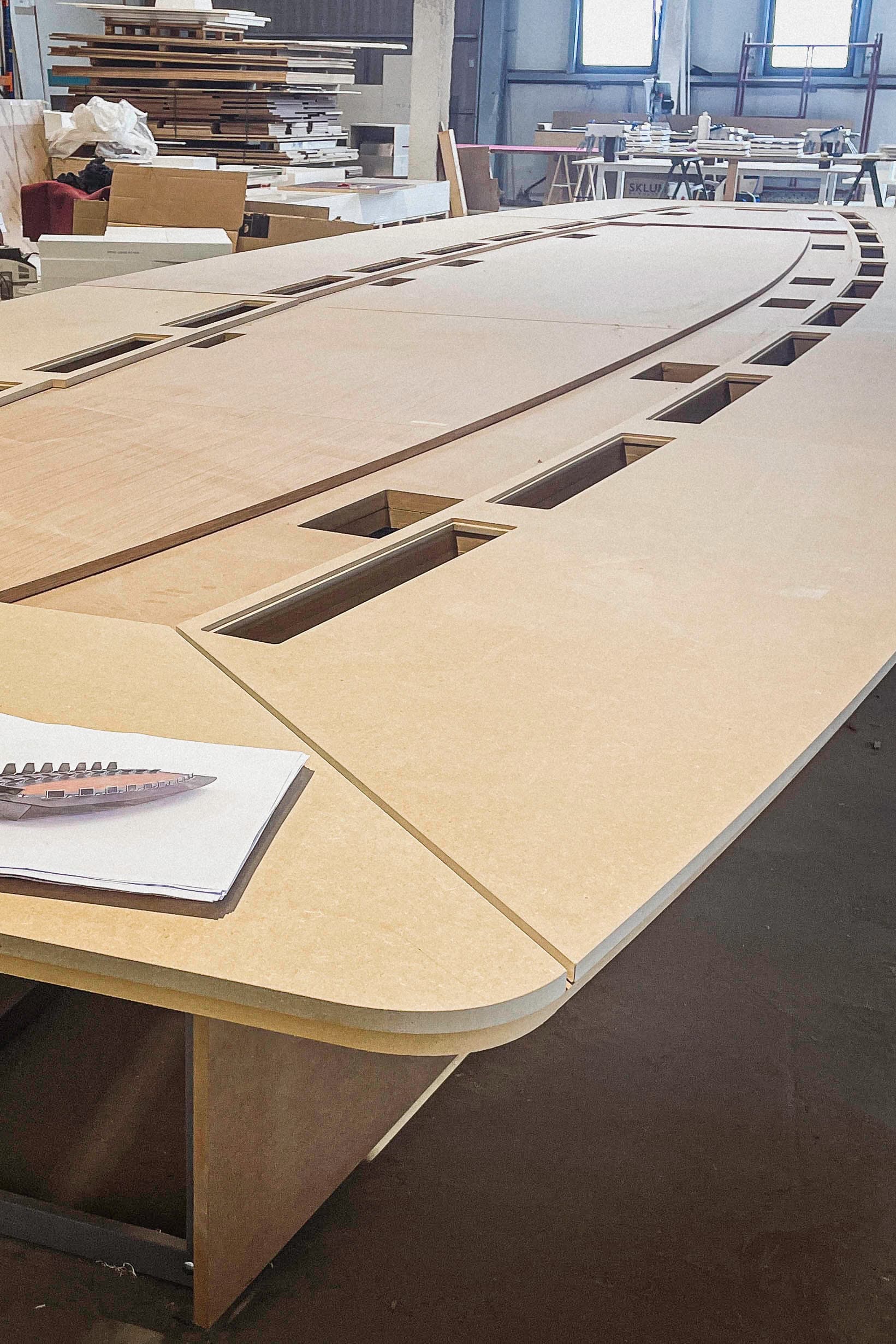 Large curved wooden structure in progress on a workshop table, with precisely cut openings and layered panels.